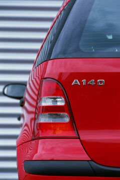 Mercedes-Benz A 140, model series 168, 2001 version, jupiter red (589), slate grey fabric (388). In the CLASSIC equipment line, the radiator louvres, rubbing strips, door handles and exterior mirror housings were in black. Special equipment: Lamella sliding sunroof (Code 417), 16-inch light-alloy wheels in 5-hole design (650). Close-up of model badge on left of tailgate.