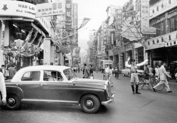 Straßenbild in  Hongkong: Hauptgeschäftsstraße in Hongkong. Ein Mercedes-Benz-Fahrzeug, Typ  180, passiert eine Kreuzung. Für einen Europäer, der eben erst in Hongkong angekommen ist, ein ungewohntes Bild, 1959.